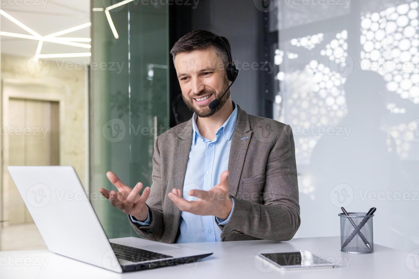 Smiling young man sitting at desk in office wearing headset and talking gesturing hands on call via laptop. photo
