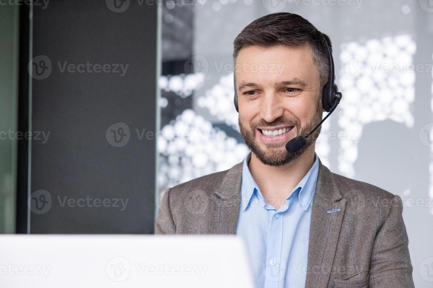 Businessman in business suit with headset phone for call smiling typing on laptop keyboard, online customer service worker at workplace inside office. photo