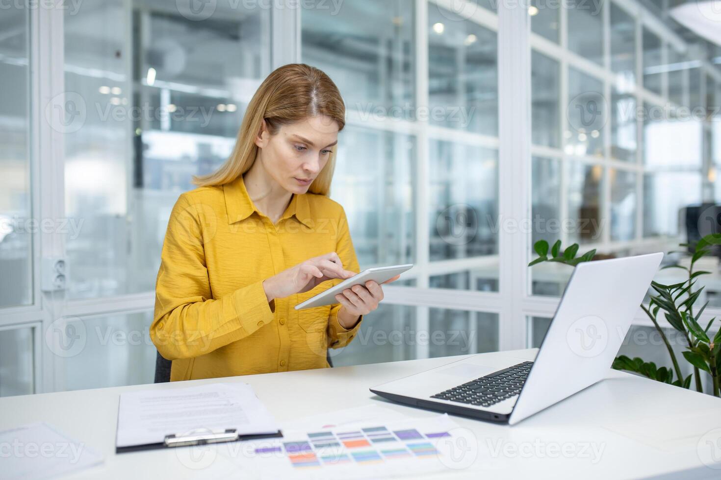 Businesswoman in yellow shirt working in modern office, using tablet and laptop, demonstrating focus and productivity. photo