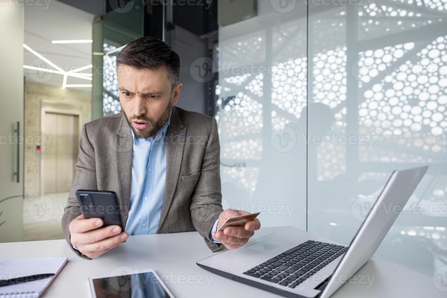 Businessman shocked by fraud notification while holding credit card and phone in office setting with laptop on desk photo