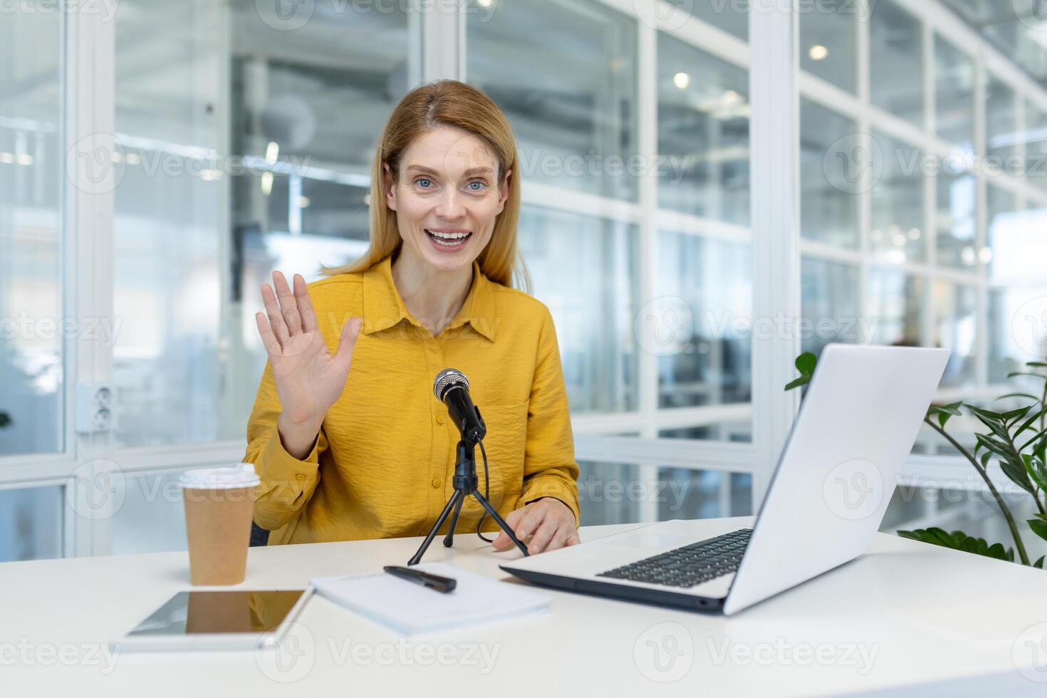 Smiling woman in a yellow shirt hosting a podcast from her office desktop, featuring a laptop and a microphone. Professional setup for online content creation. photo