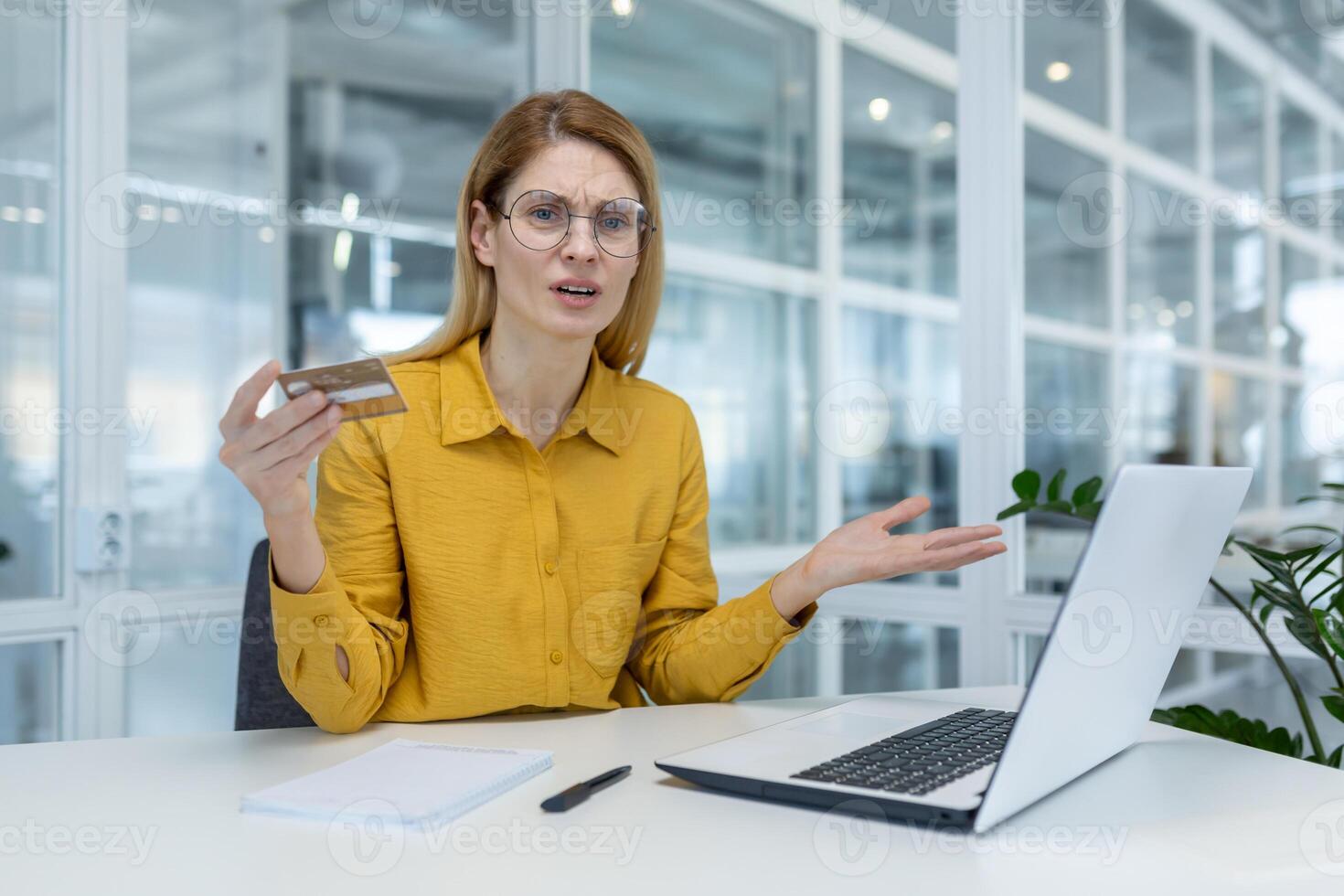 Concerned woman holding credit card while using laptop in office. She appears frustrated, likely dealing with an online transaction or financial issue. photo