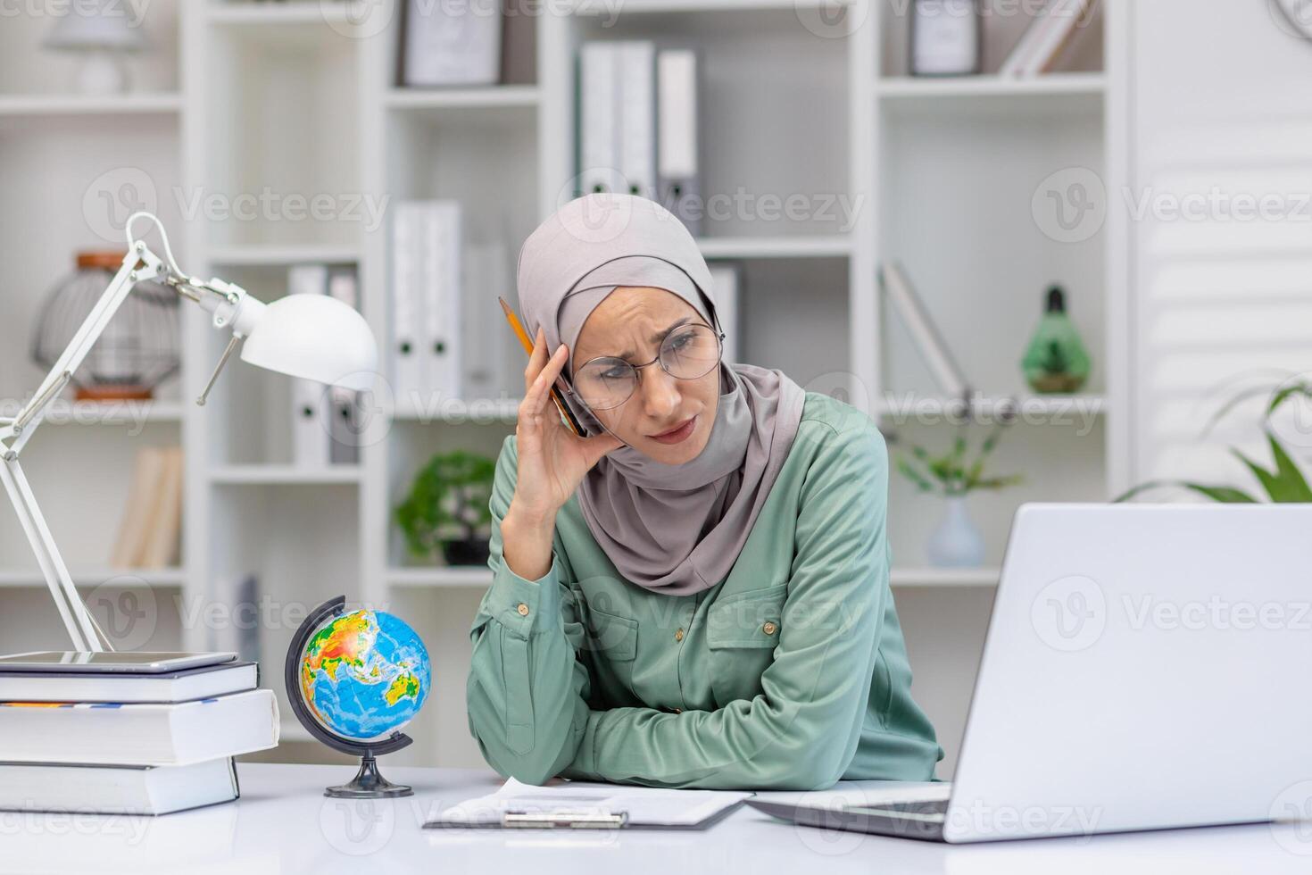 Stressed woman in hijab working on laptop at office desk with globe, books, and lamp. Concept of business, education, remote work, fatigue. photo