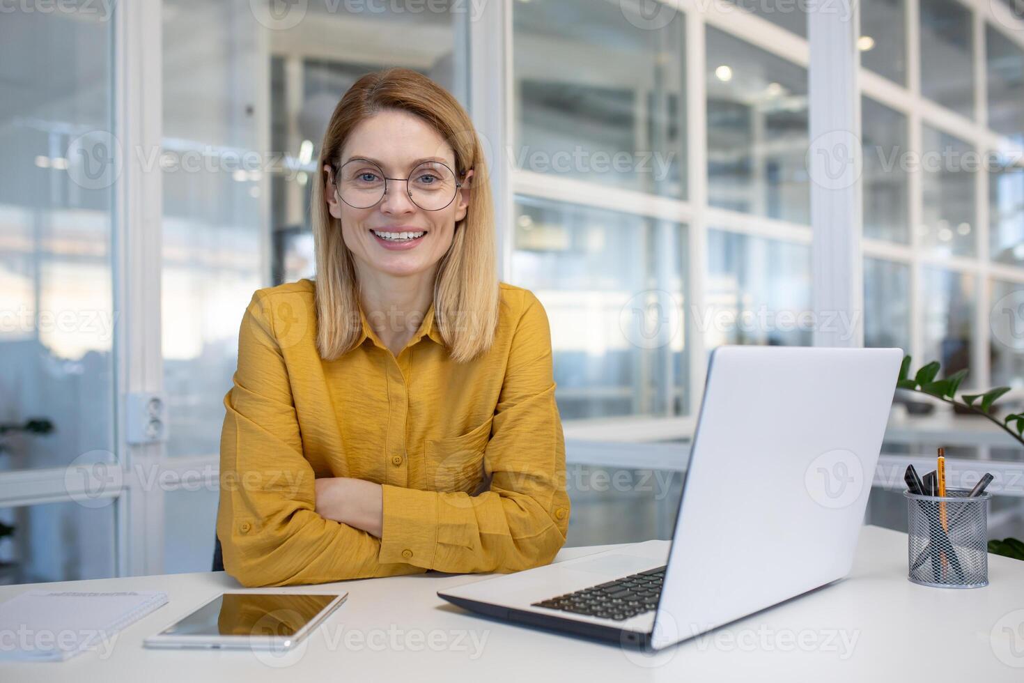 Smiling woman in a modern office setting, sitting at a desk with a laptop, wearing glasses and a yellow blouse with confidence. photo