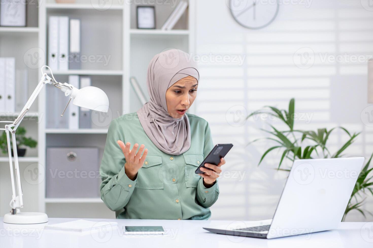 Muslim professional in hijab with shocked expression looking at smartphone, laptop and office supplies on desk. photo
