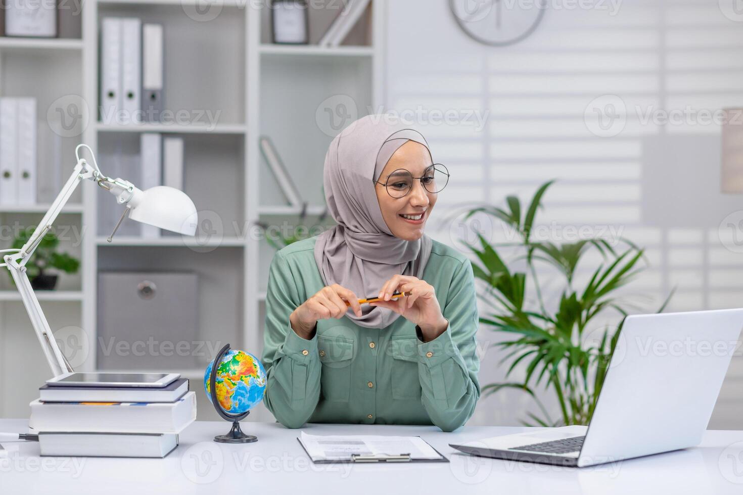 A cheerful Muslim woman wearing a hijab engaged in online teaching, using a laptop and pointing at a small globe, surrounded by books in a well-organized office. photo