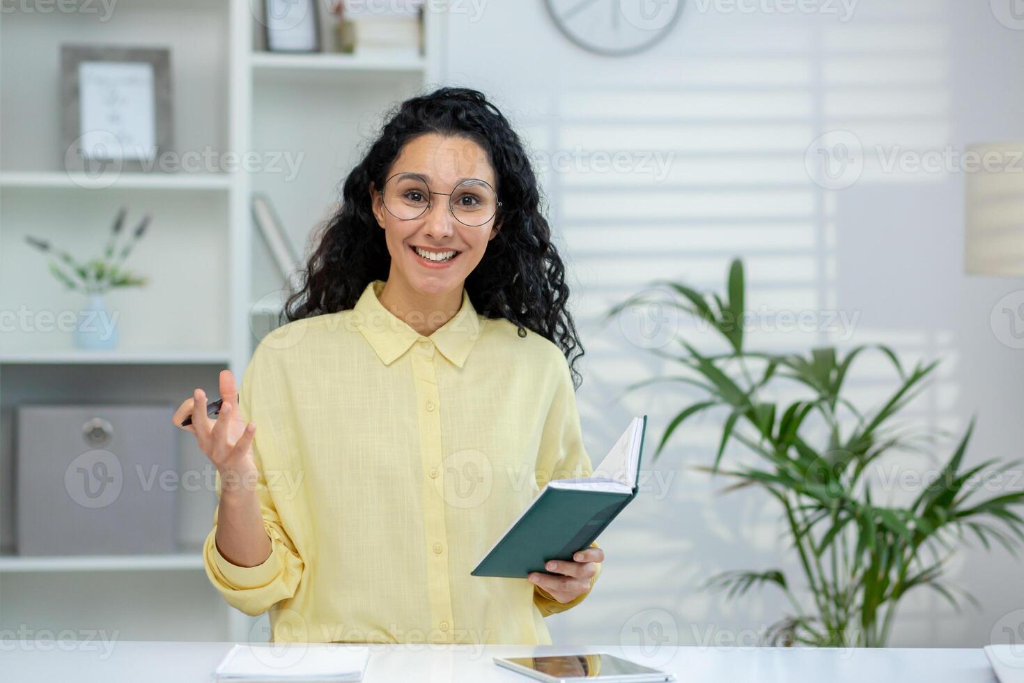 A smiling Hispanic woman conducting an online tutorial with a student, using gestures and holding a notebook in a bright office setting. photo
