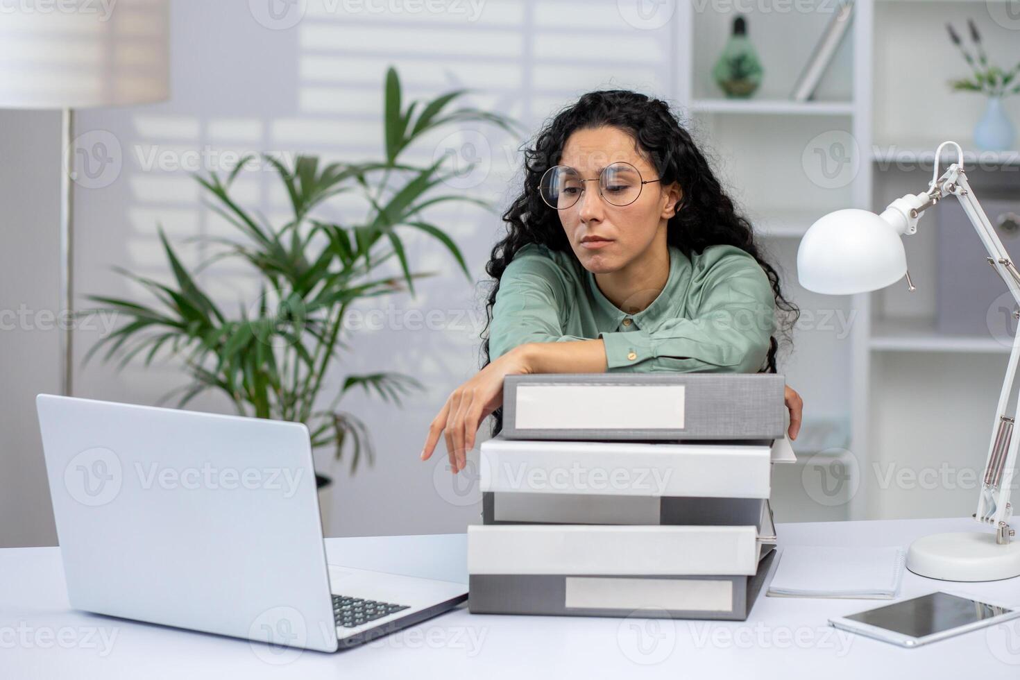 A Hispanic woman in an office environment, leaning on a stack of books, appears stressed and overwhelmed while staring at a laptop screen. photo