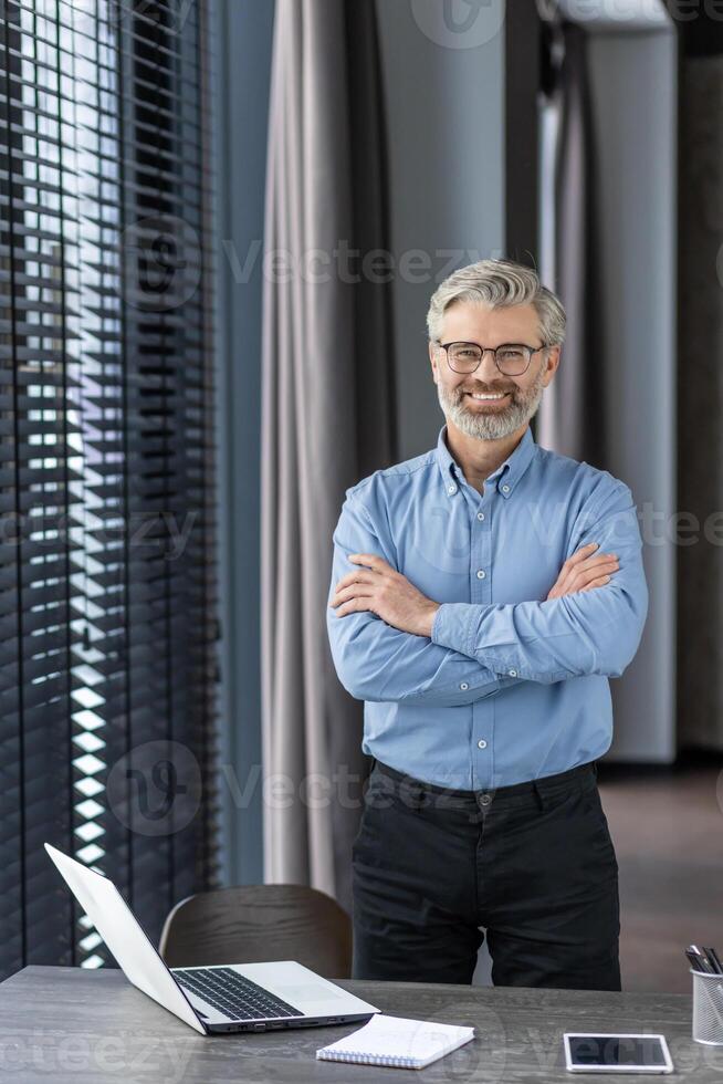 Confident mature businessman with glasses standing in a modern office, arms crossed, smiling at the camera, with a laptop and phone on desk. photo