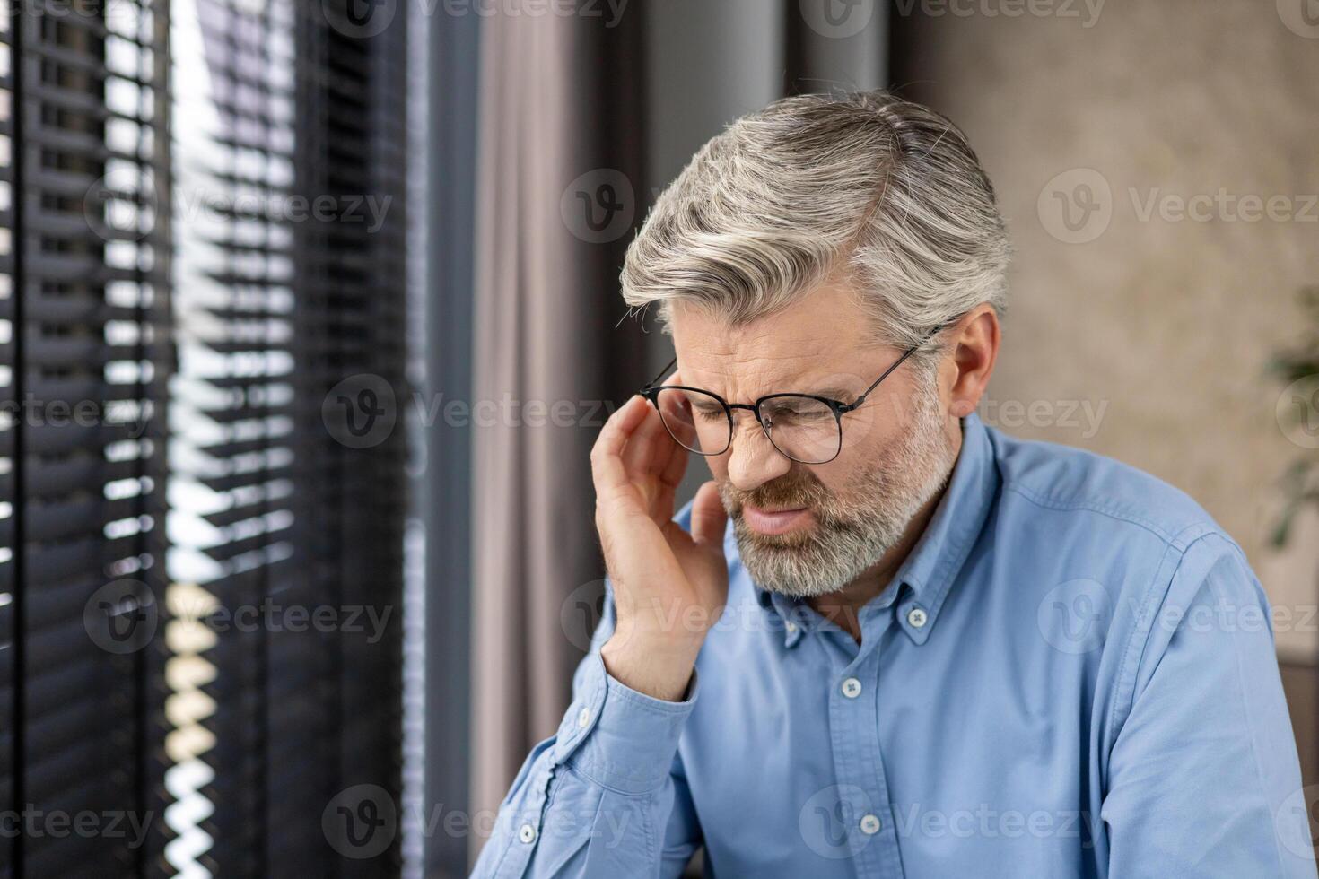 Elderly man with glasses and grey hair deep in thought by a window with blinds, in a calm contemplative mood photo