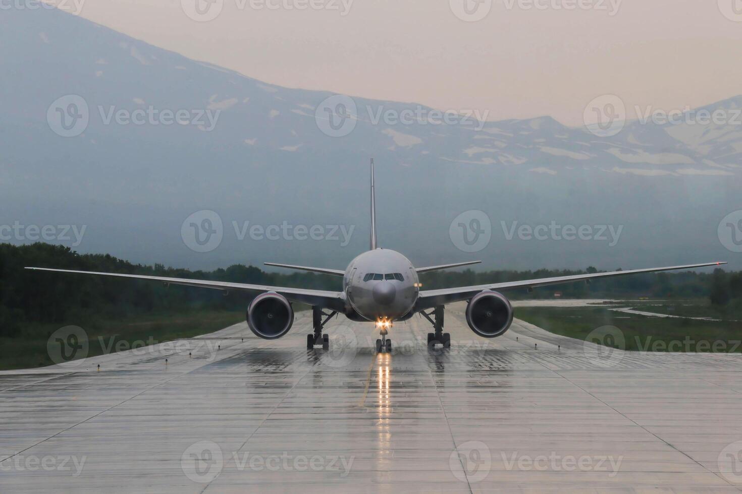 Frontal view of Aircraft coming in for Landing against background of Snow-capped Mountain Peaks on Rainy cloudy day. The Light Path from Headlights creeps along the Wet runway towards the airport photo