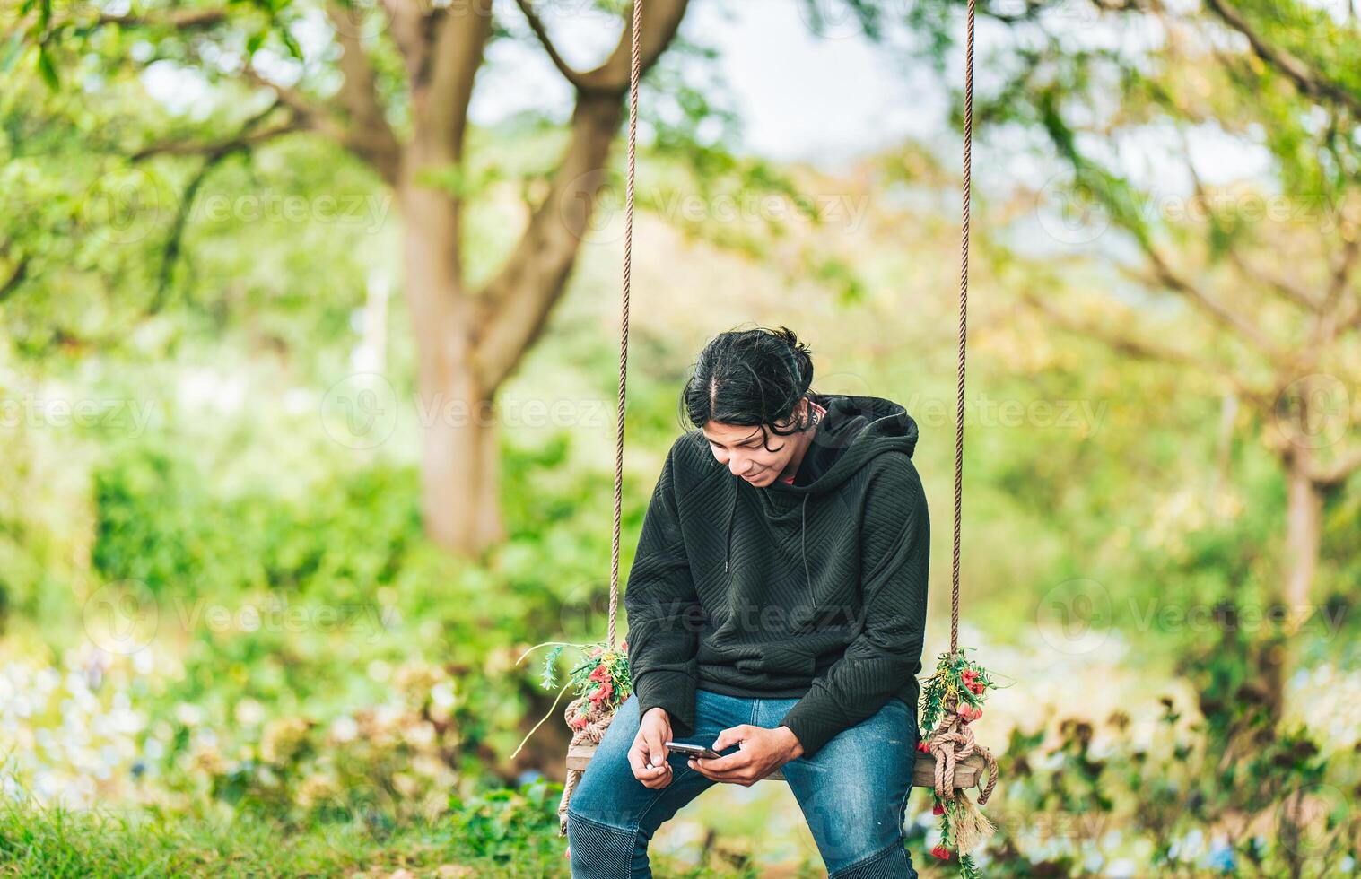 Smiling guy sitting on a swing with smart phone. Young man sitting on a swing using cell phone in nature photo