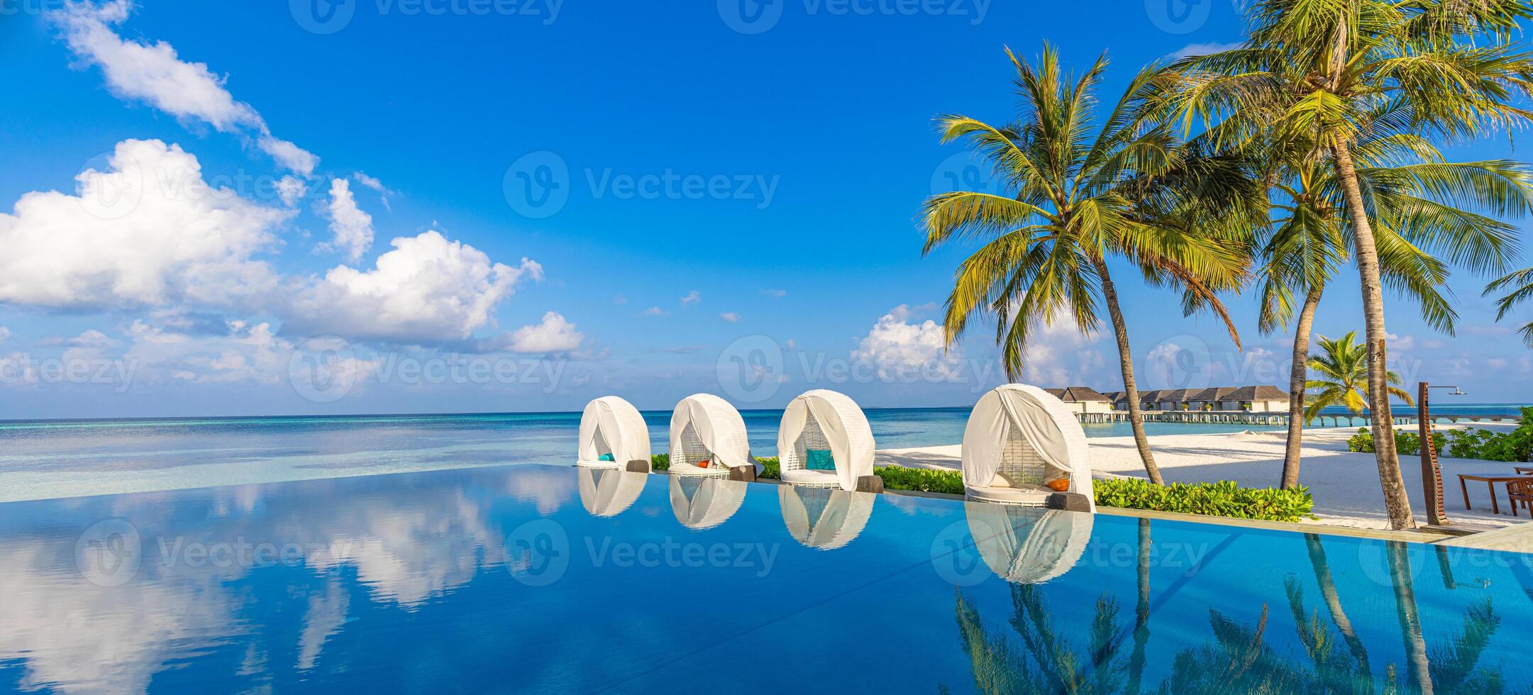 Outdoor tourism landscape. Luxurious beach resort with swimming pool and beach chairs or loungers umbrellas with palm trees and blue sky, sea horizon. Summer island relax travel and idyllic vacation photo