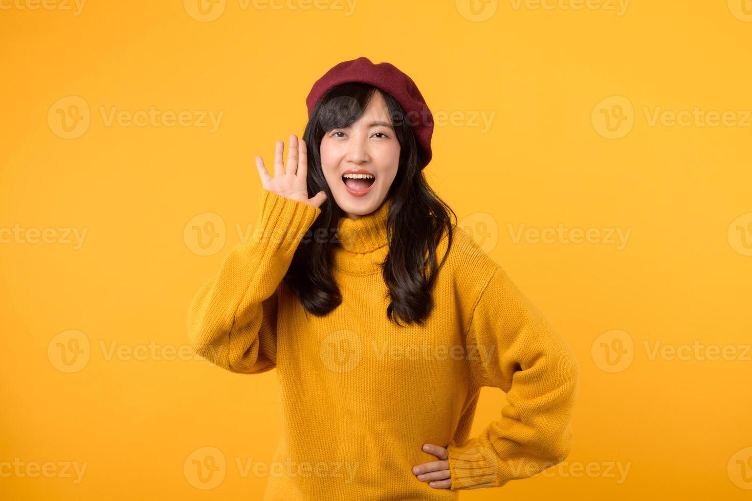 Confident young Asian woman in her 30s making a fashionable announcement, adorned in a yellow sweater and red beret against a vibrant yellow backdrop. photo