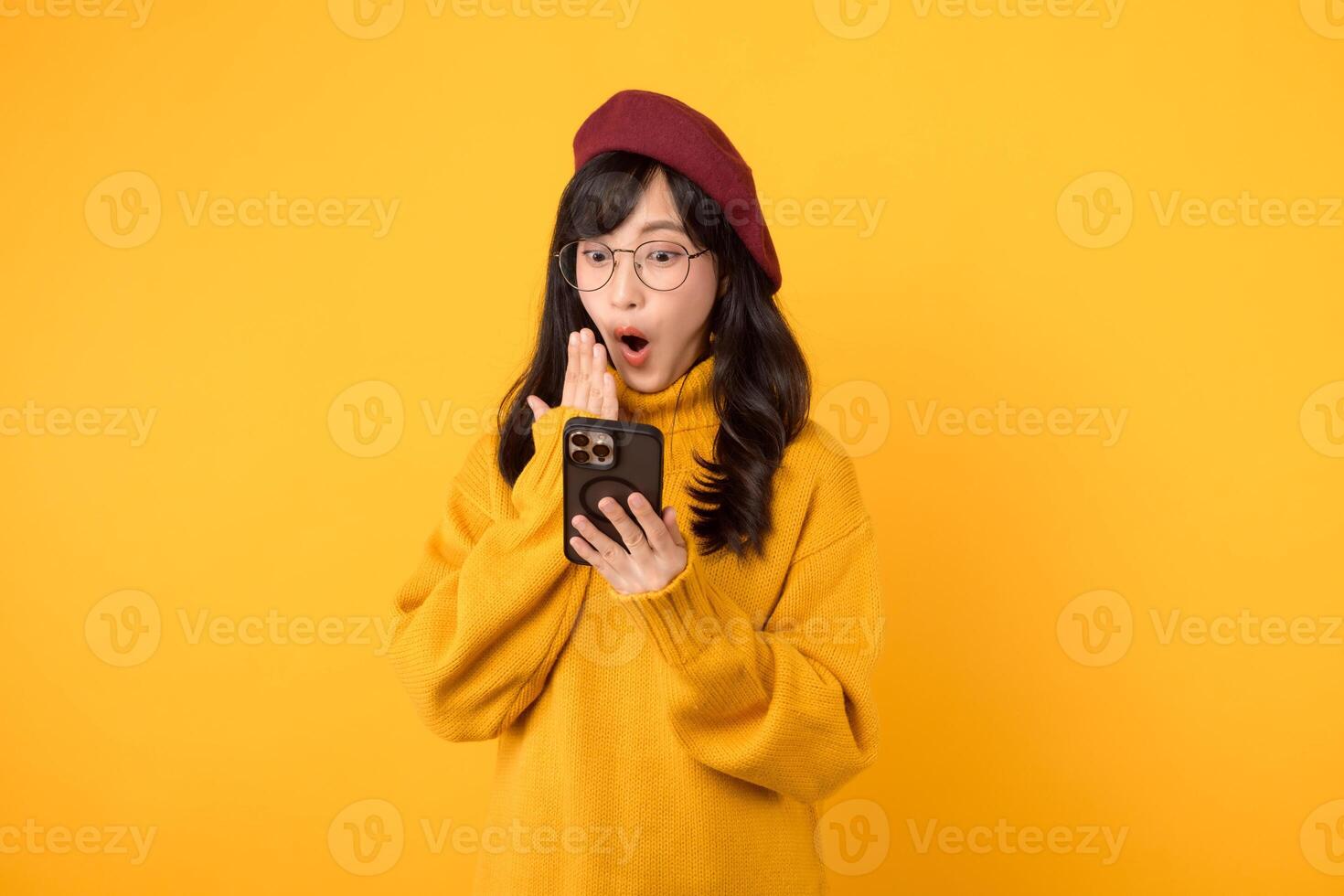 Excited young woman using smartphone, wearing yellow sweater, red beret, and eyeglasses against a vibrant yellow background. photo