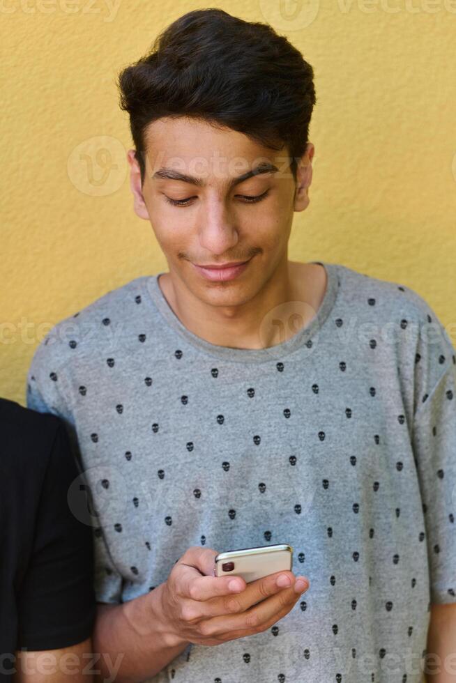 Middle Eastern Teenager Using Mobile Phone While Leaning Against Yellow Wall photo