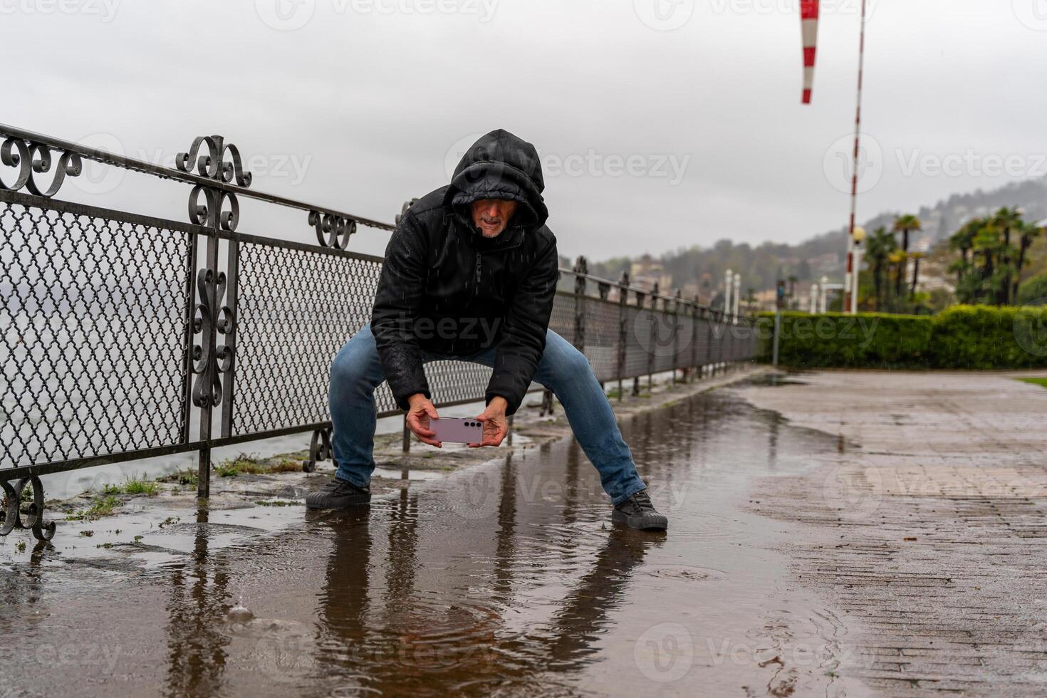 middle aged hipster man photographing a puddle with mobile phone on the lakeside in the pouring rain photo