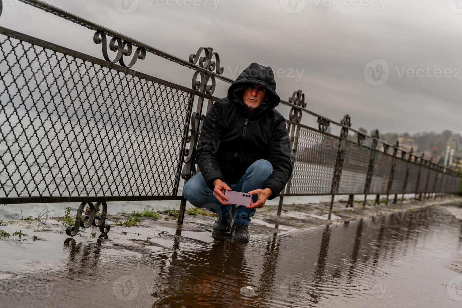 middle aged hipster man photographing a puddle with mobile phone on the lakeside in the pouring rain photo