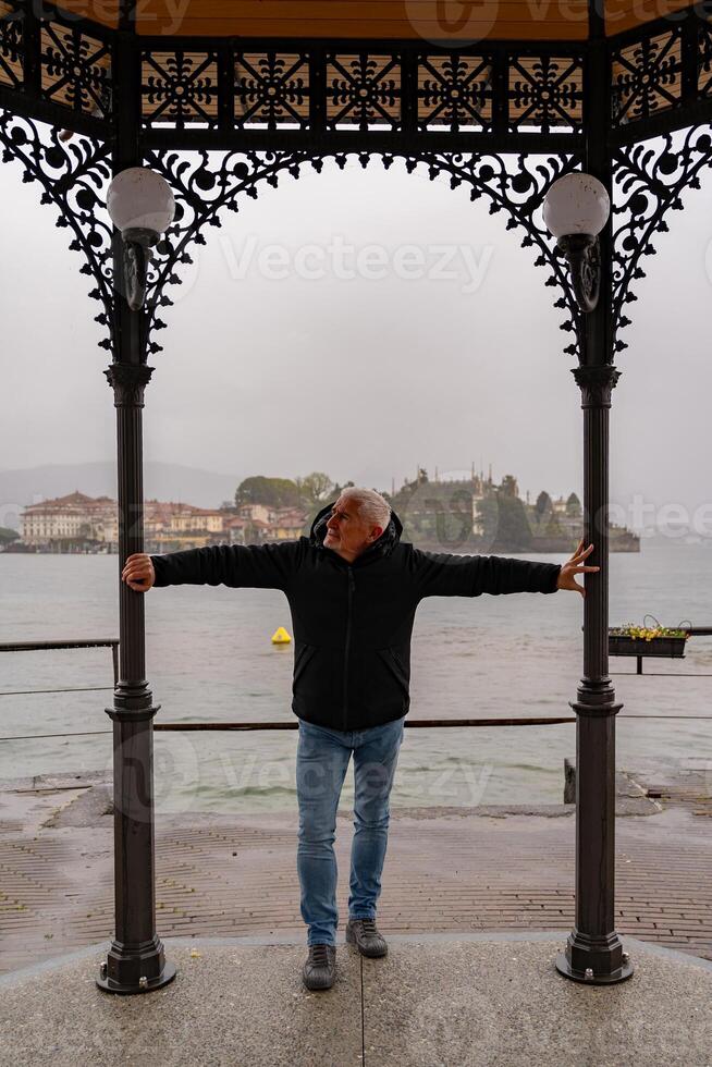 middle aged man wearing a black windbreaker posing under a gazebo by the lake on a rainy day - travel and tourism concept photo