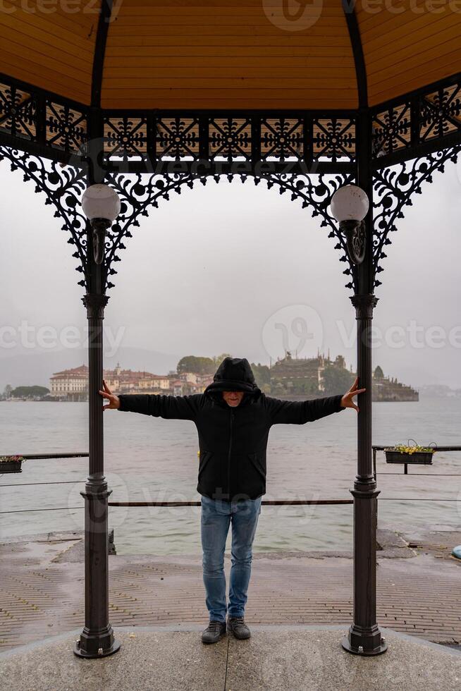 middle aged man wearing a black windbreaker posing under a gazebo by the lake on a rainy day - travel and tourism concept photo