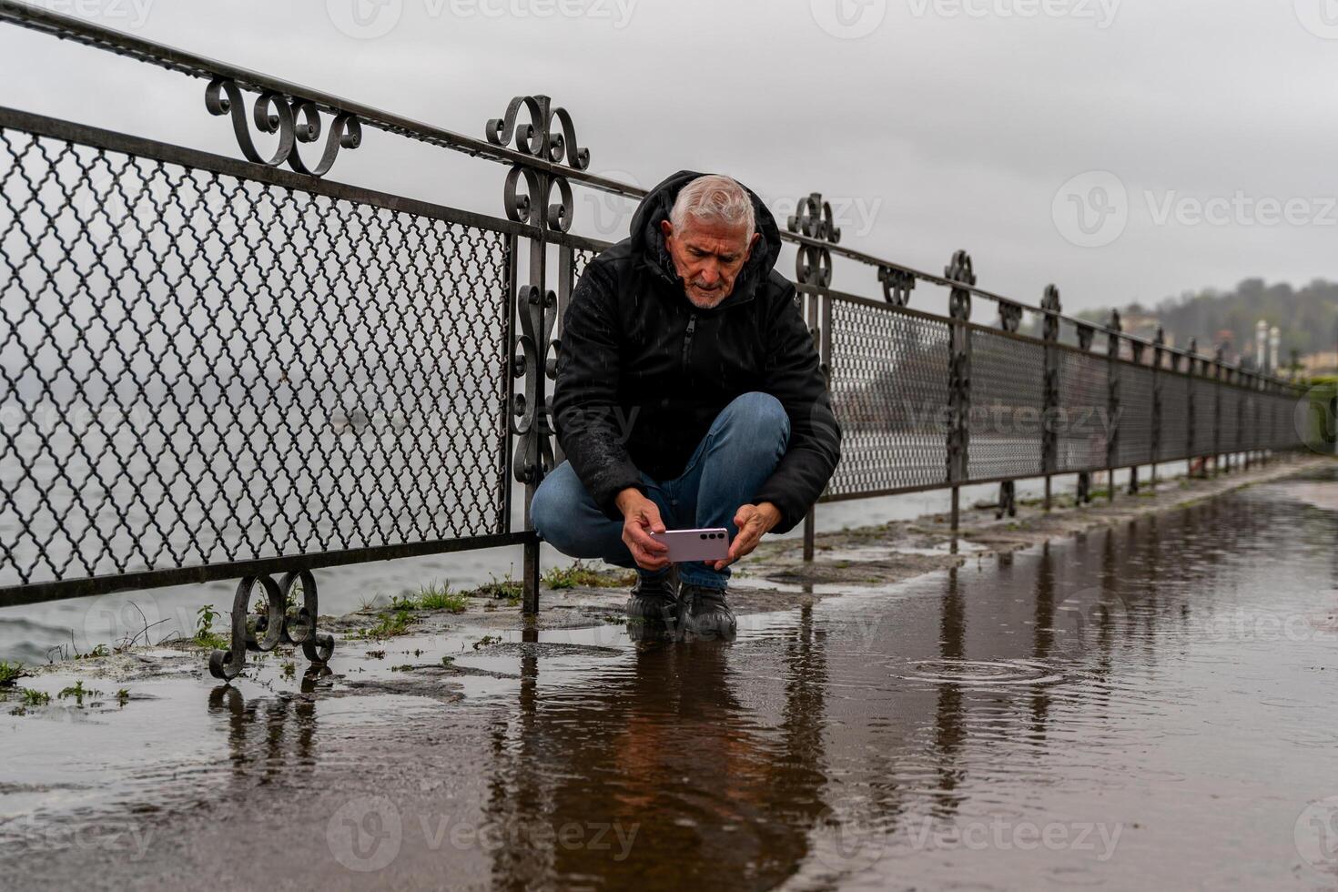 middle aged hipster man photographing a puddle with mobile phone on the lakeside in the pouring rain photo