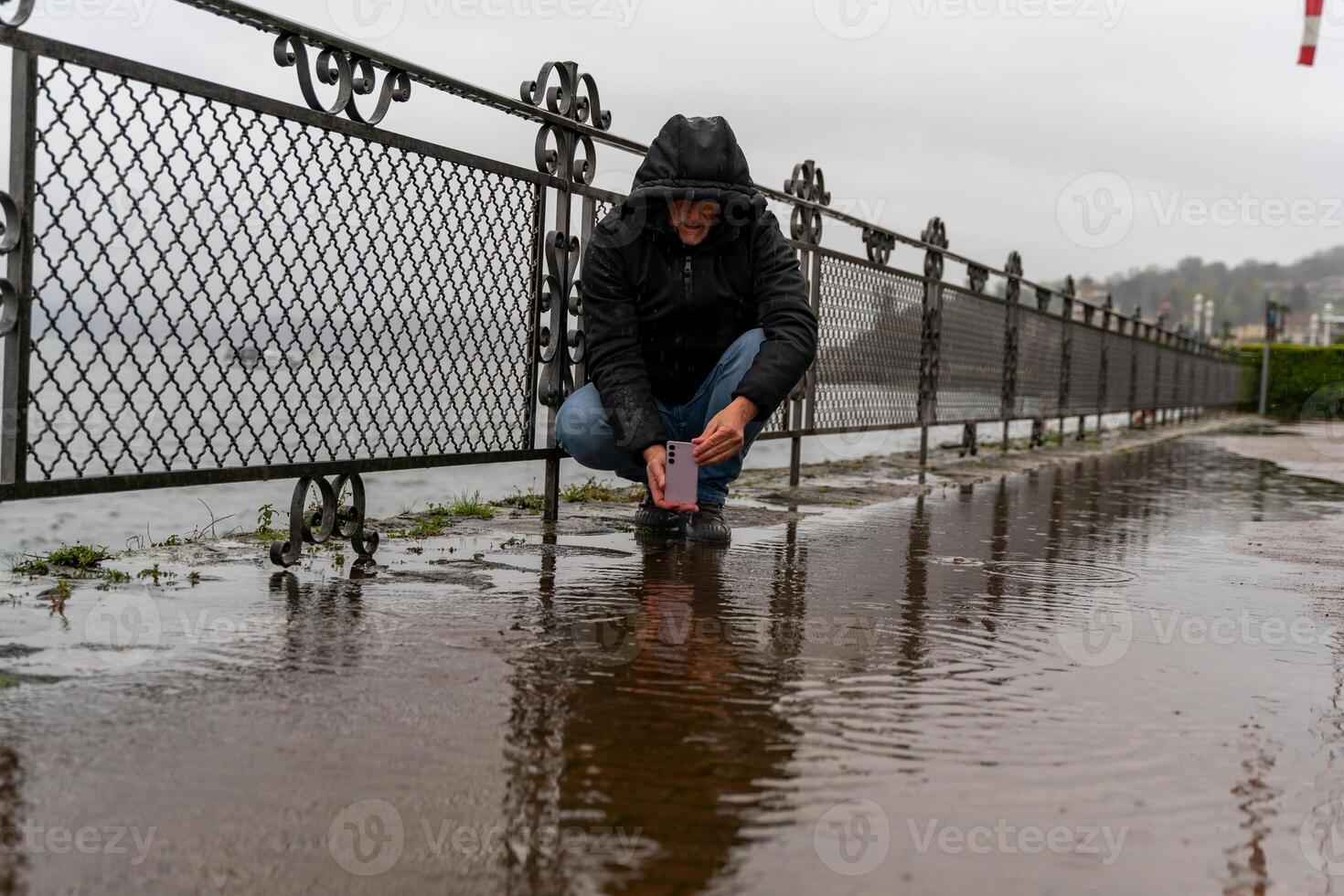 middle aged hipster man photographing a puddle with mobile phone on the lakeside in the pouring rain photo