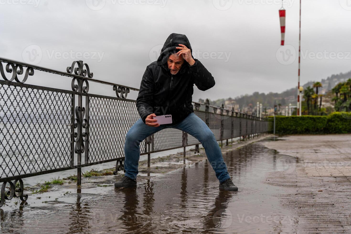 middle aged hipster man photographing a puddle with mobile phone on the lakeside in the pouring rain photo
