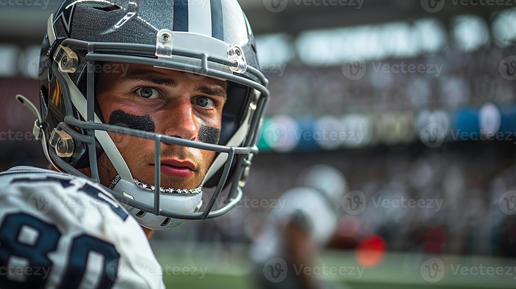 American football player wearing a gray helmet and a white uniform with black stripes under his eyes. . There is a football field and stands with fans in the background. Close-up of the face photo