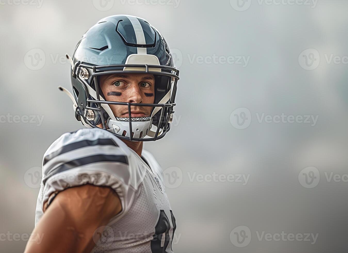 American football player in a gray helmet and white uniform with black stripes under his eyes, he stands sideways looking to the right. Copy space photo
