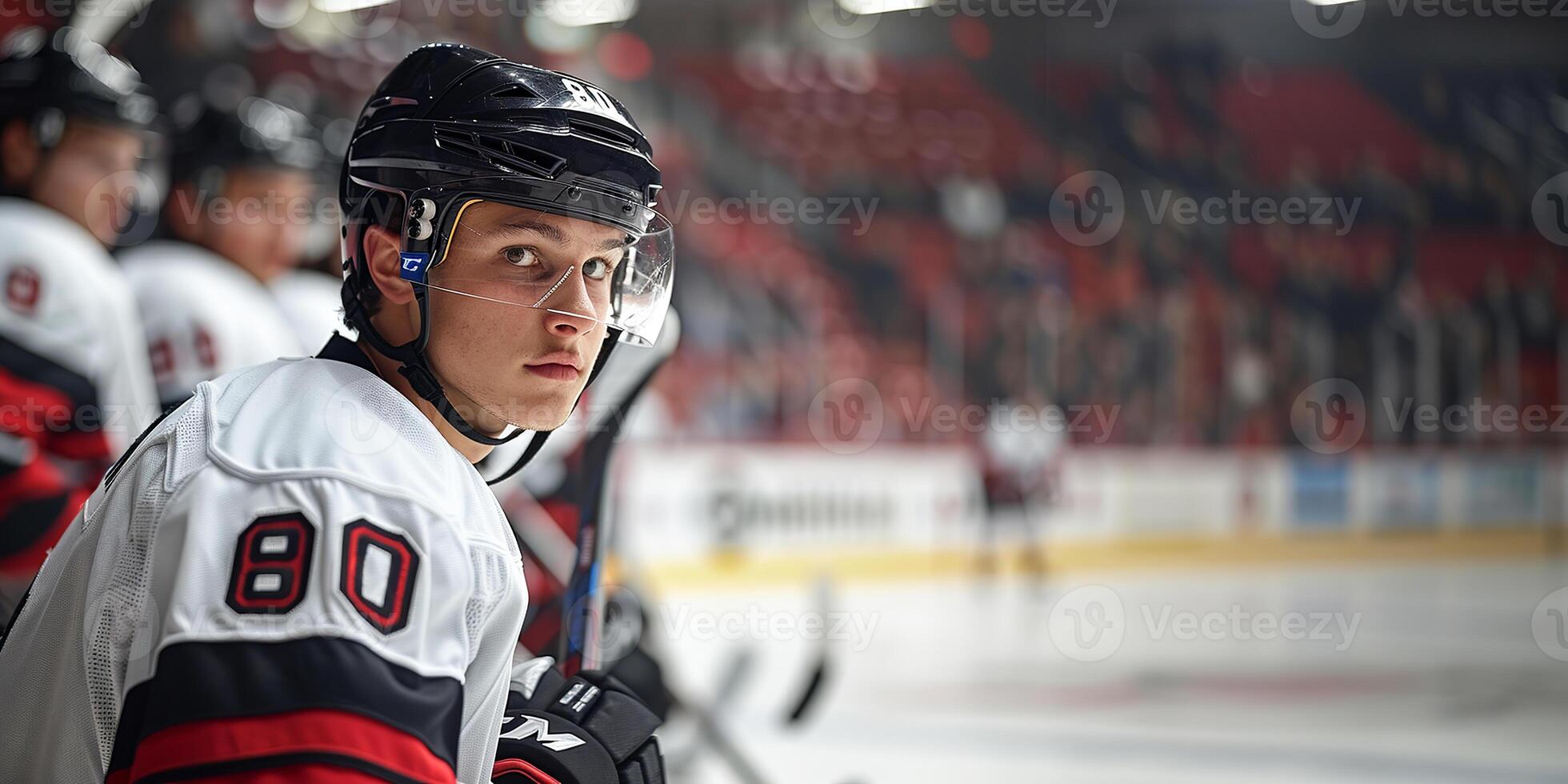 A hockey player in a black helmet and white jersey sits on the bench. Hockey arena in the background. Copy space photo
