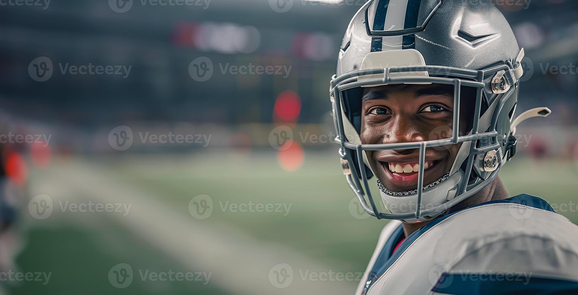 American football player in a black helmet and white uniform smiles and gives a thumbs up in the right part of the frame. There is a football field and stands with fans in the background. Copy space photo