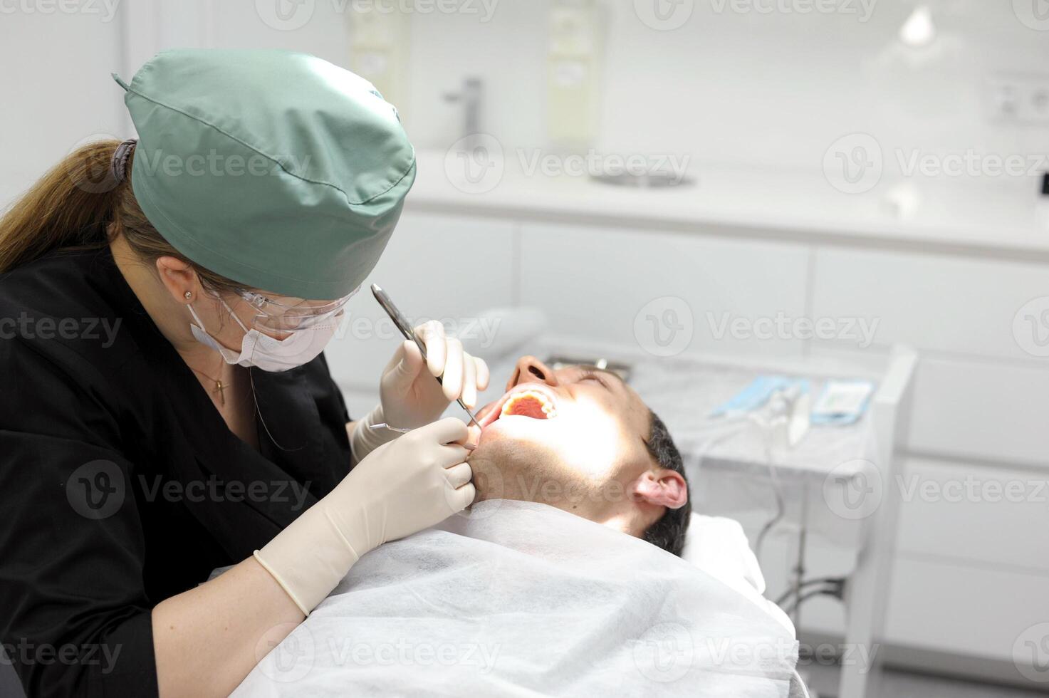 Young female dentist drilling tooth to patient female dentist green hat black clothes treats male patient operation surgical gloves pull out tooth extract photo