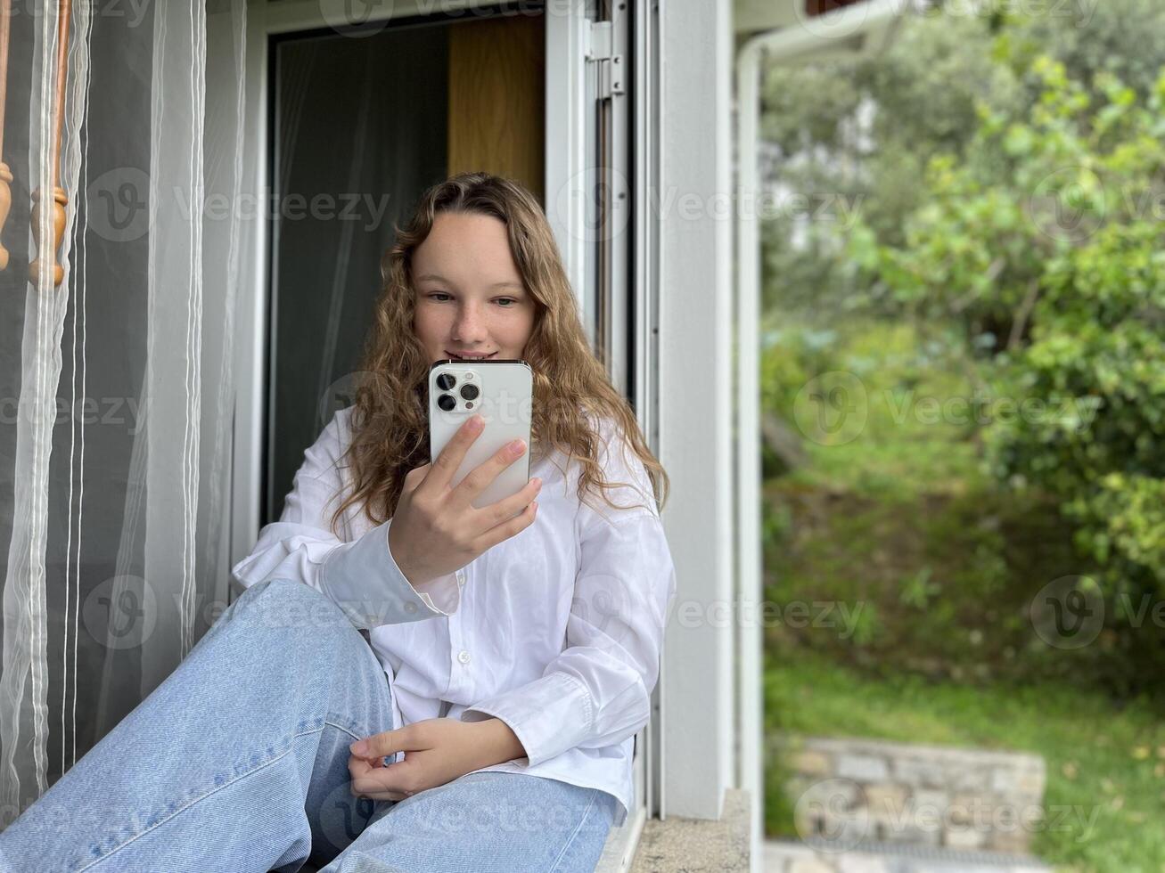 a girl in a white shirt a teenager sits on a white windowsill the view from the street she can be seen in the window in her hands she holds a white iPhone 13 pro max she looks at the screen calmly photo