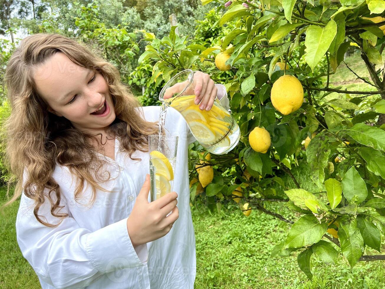 it's just a lemon paradise, a teenage girl stands against the backdrop of a lemon tree with lemons and drinks lemonade from a glass glass in her other hand, she holds a jug of drink photo