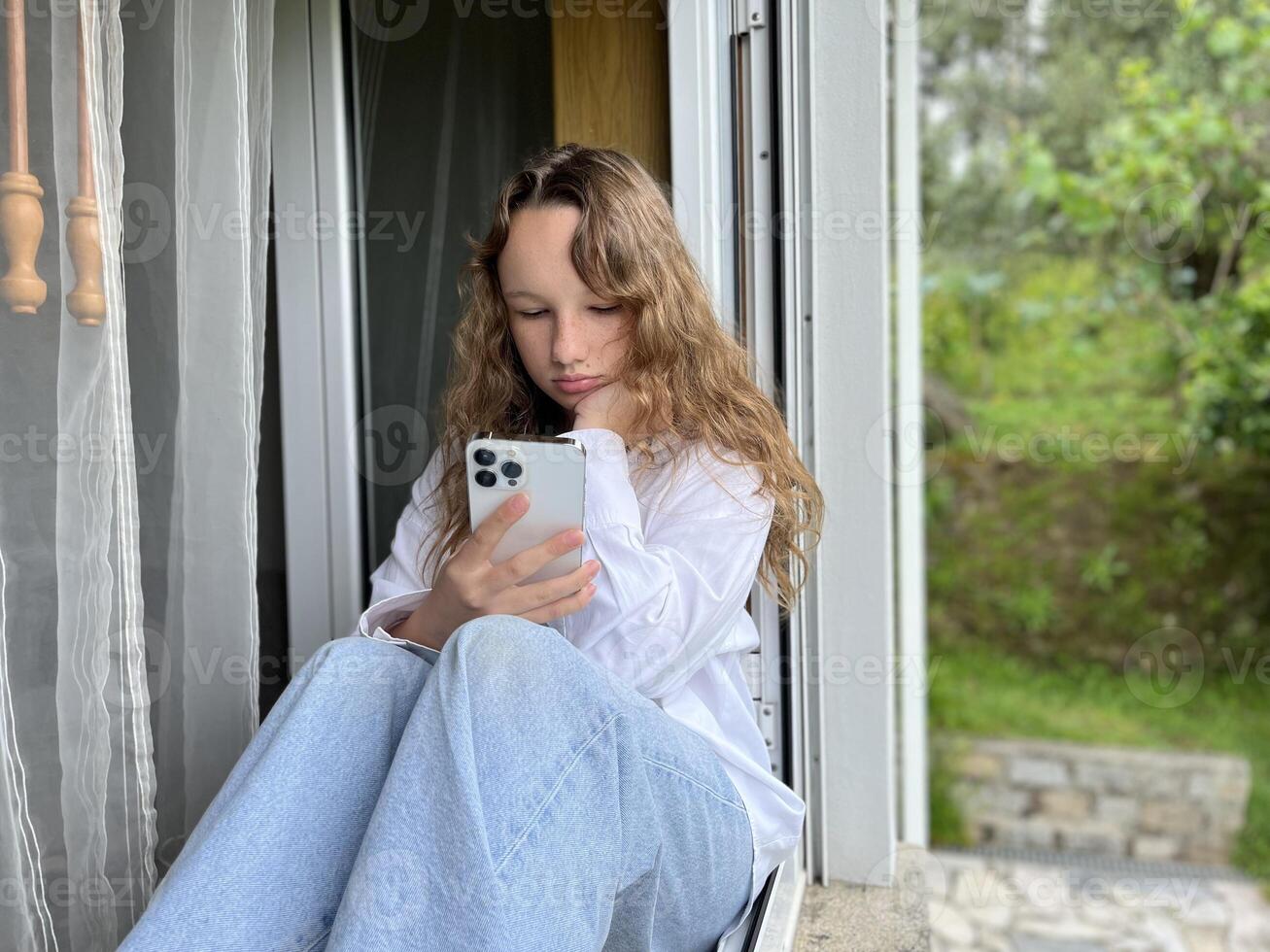 a girl in a white shirt a teenager sits on a white windowsill the view from the street she can be seen in the window in her hands she holds a white iPhone 13 pro max she looks at the screen calmly photo