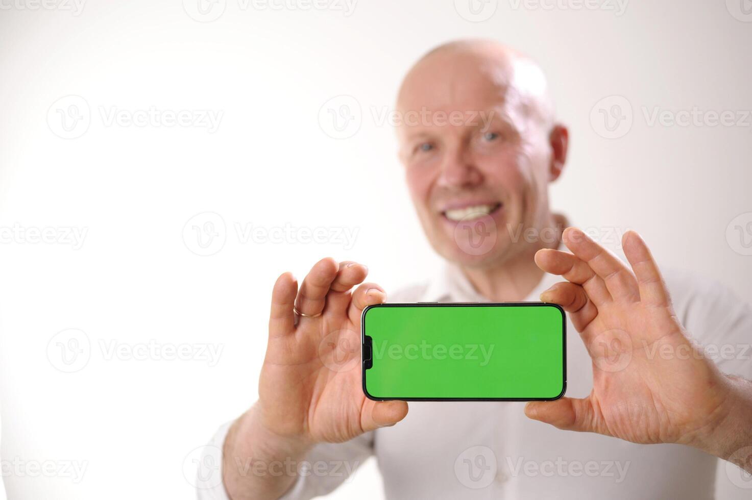 Portrait of positive funky aged person indicate finger empty space proposition shows off something on smartphone with green screen to camera. Focusing on smartphone with chromakey photo