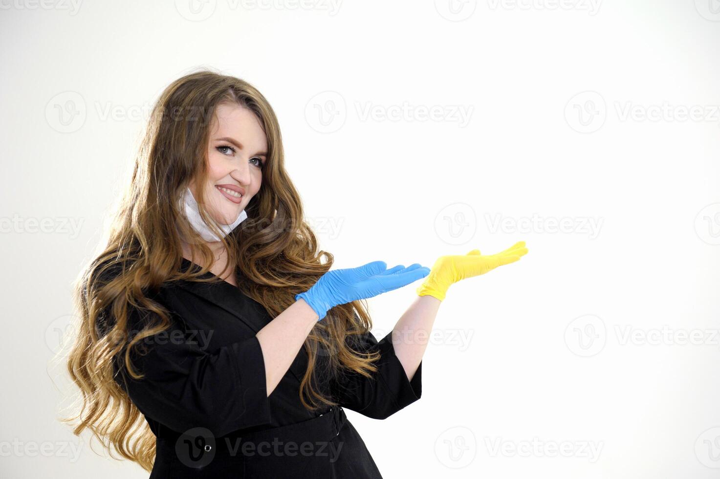a female doctor in a black suit with flowing hair with a mask lowered to her chin points with her hand to an empty space for advertising text blue and yellow hygienic glove studio white background photo