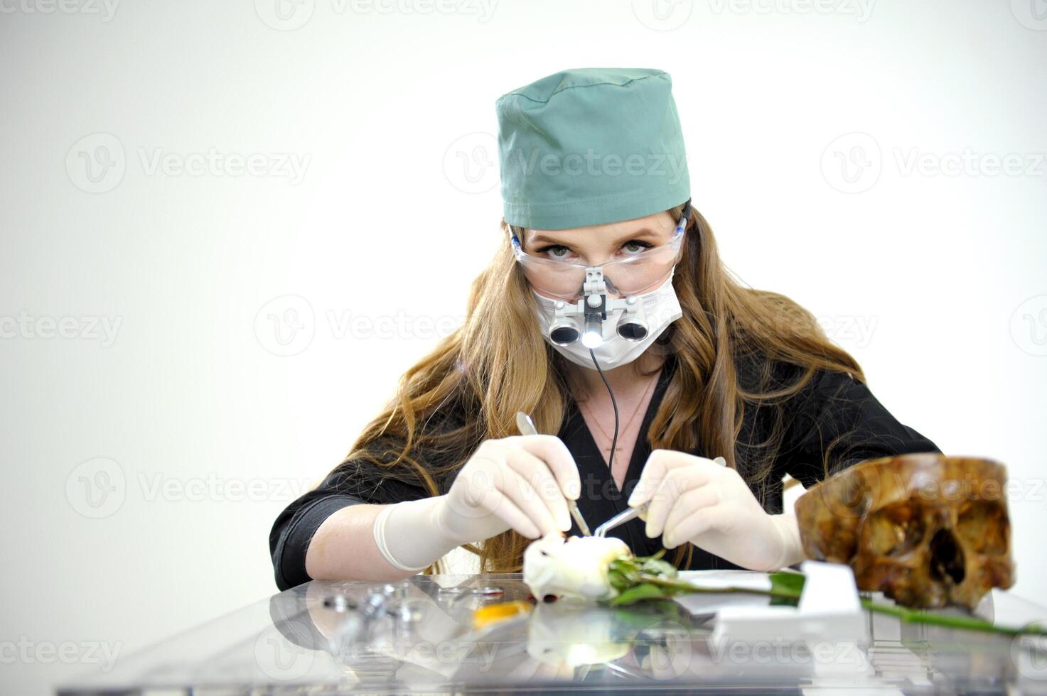 Illuminator with safety glasses and binoculars woman doing surgery showing demonstrating on a flower with scalpel next to skull blank text surgical latex gloves black clothes doctor's green cap photo