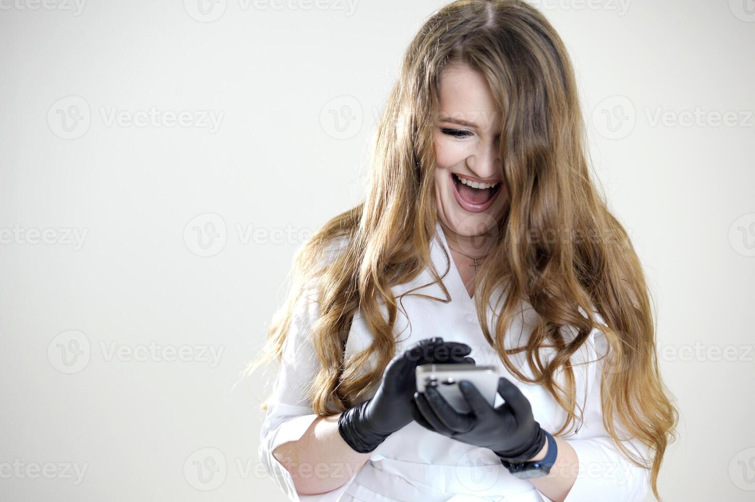 happy contented cheerful woman doctor workwear white laughs open mouth looks into mobile phone learns news latest technology communication online conference meeting with friends and gorgeous hair photo