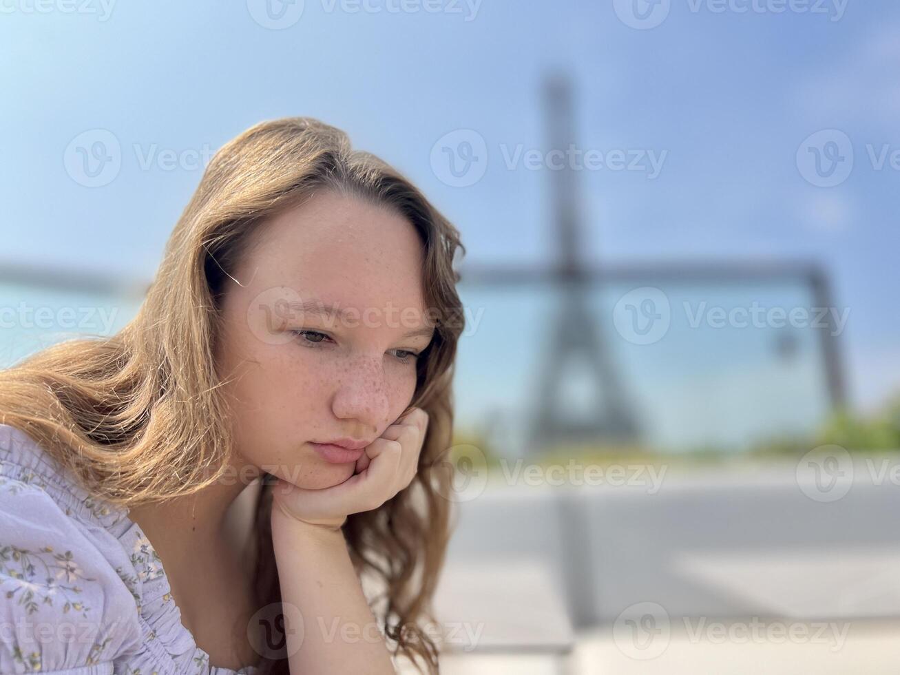 the face of a beautiful girl she is sad leaning on her arm in the distance you can see the Eiffel Tower the girl is young she is a teenager photo