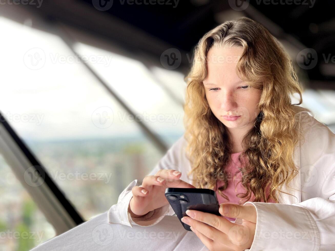 a teenage girl in a white shirt with blond hair sits and looks at the iPhone 13 phone she is interested And with her finger something corrects the black phone Top Of Vancouver Revolving Restaurant photo