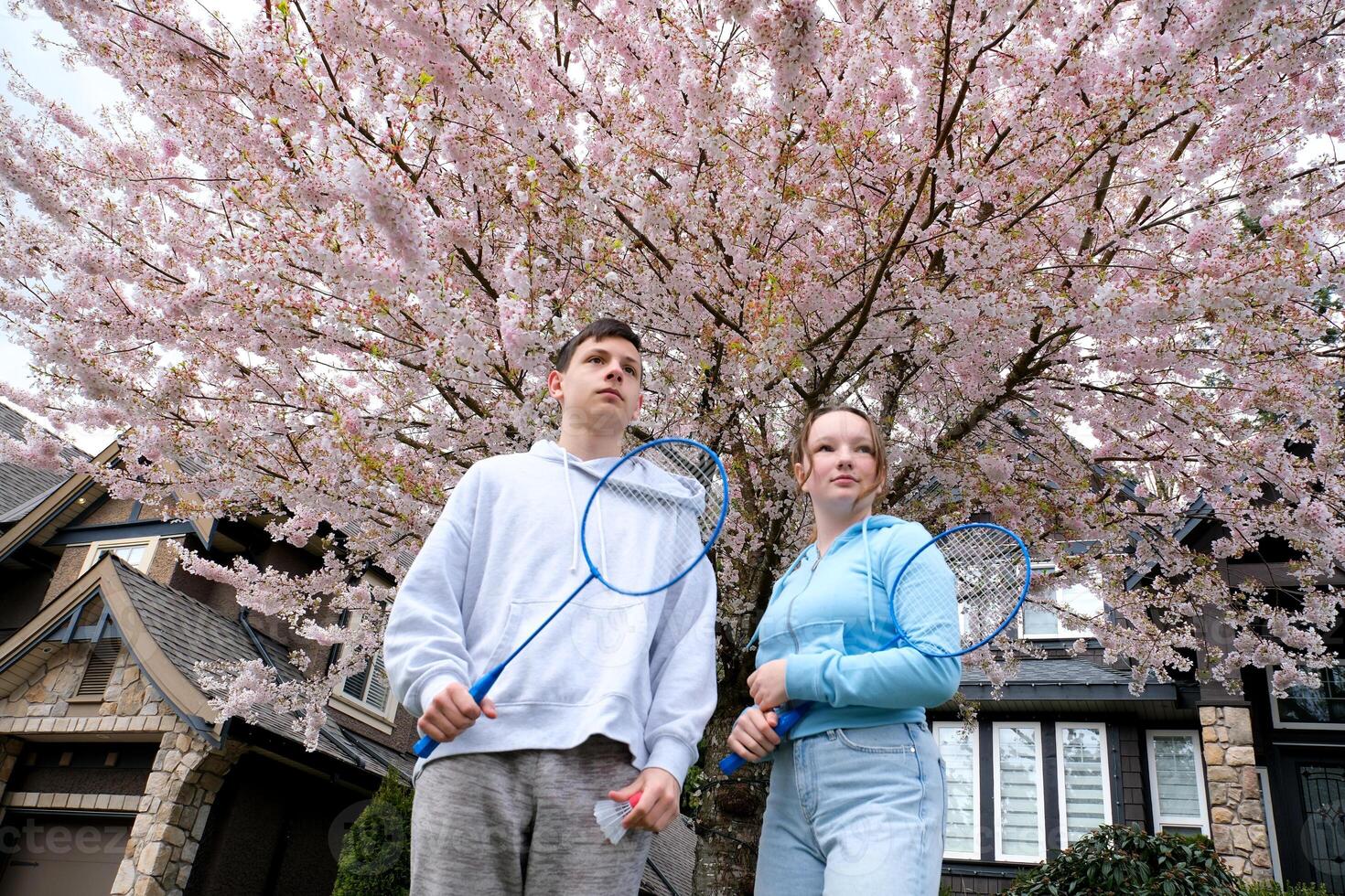 couple with badminton racquet standing together in park cherry blossoms in Surrey children stand rackets look to side adolescence spend time together at weekend sports games boy and girl teenagers photo