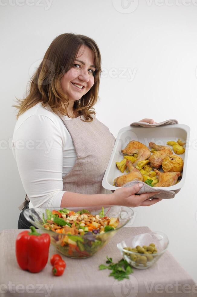 adult beautiful plump woman preparing dinner on baking sheet chicken legs quarter bowl with salad nuts ingredients smiles looks shows delicious healthy food diet family dinner lunch mom wife hostess photo