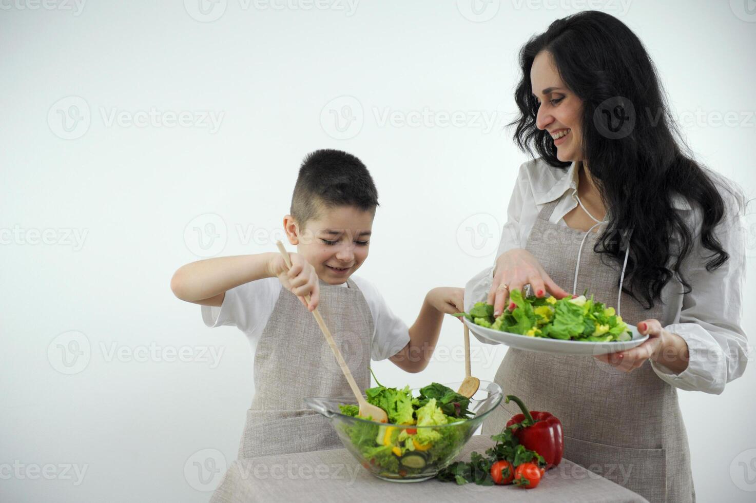 mom and son cooking fresh vegetable salad leaves smiling matching clothes apron white background studio space for text grocery store ad restaurant home food spending time with kids care love photo