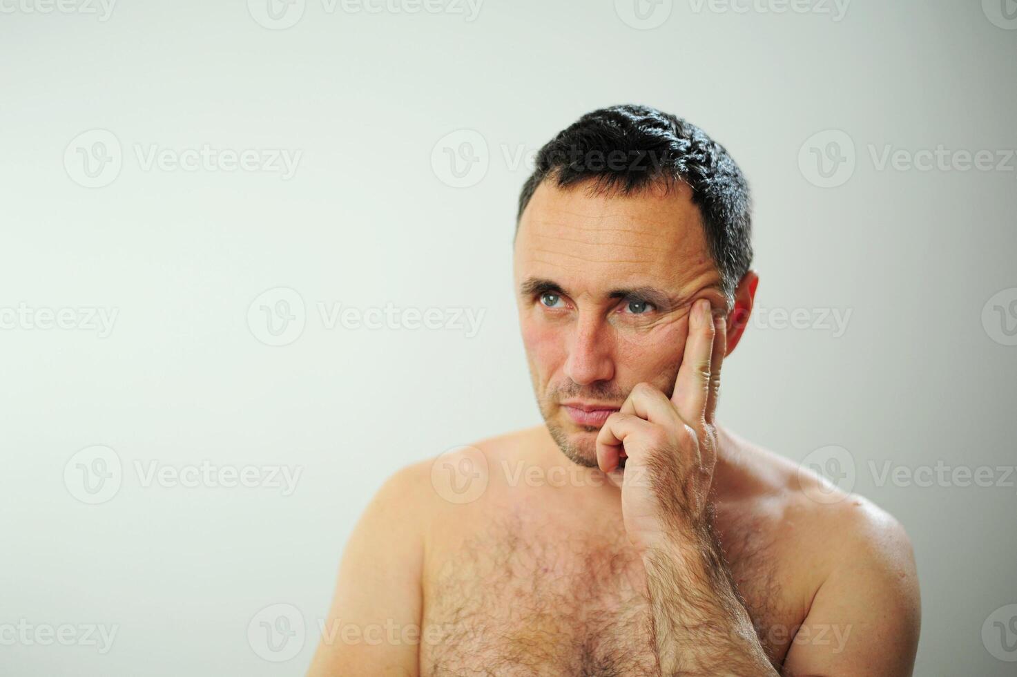 Adult man with naked torso Tired worries thinks about something He has sad tired eyes blue green color He stands on a gray background waist-deep close-up When will it end when will the best time come photo