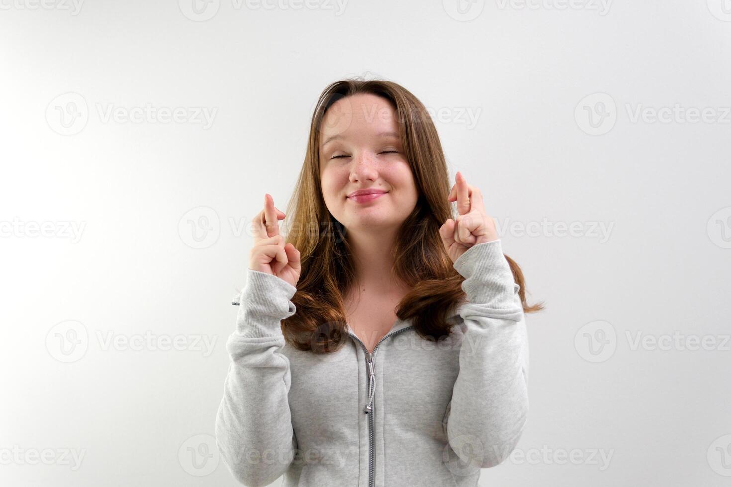 Intrigued woman in t-shirt praying with crossed fingers and looking away over grey background photo