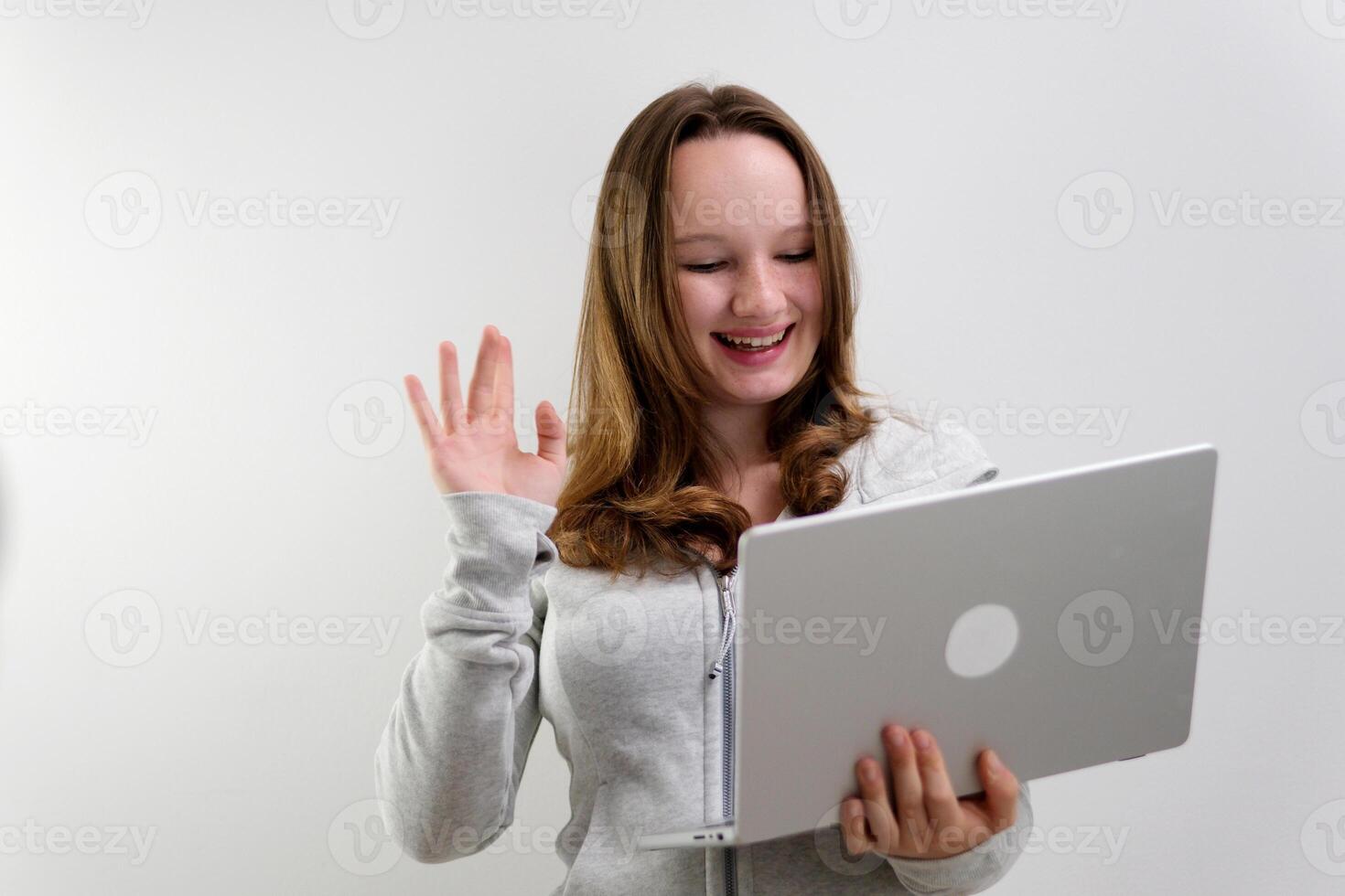 Joyful young female in wireless headphones waving wand greeting to screen while sitting at table and having chat with business partners using laptop against blurred dark interior photo
