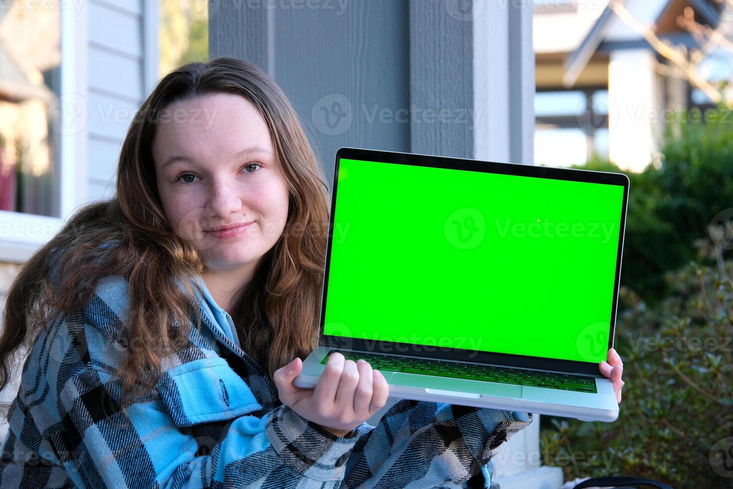 Portrait studio shot of young hipster female model in casual street wears sunglasses sit on floor holding pointing green blank empty screen laptop computer for advertising photo