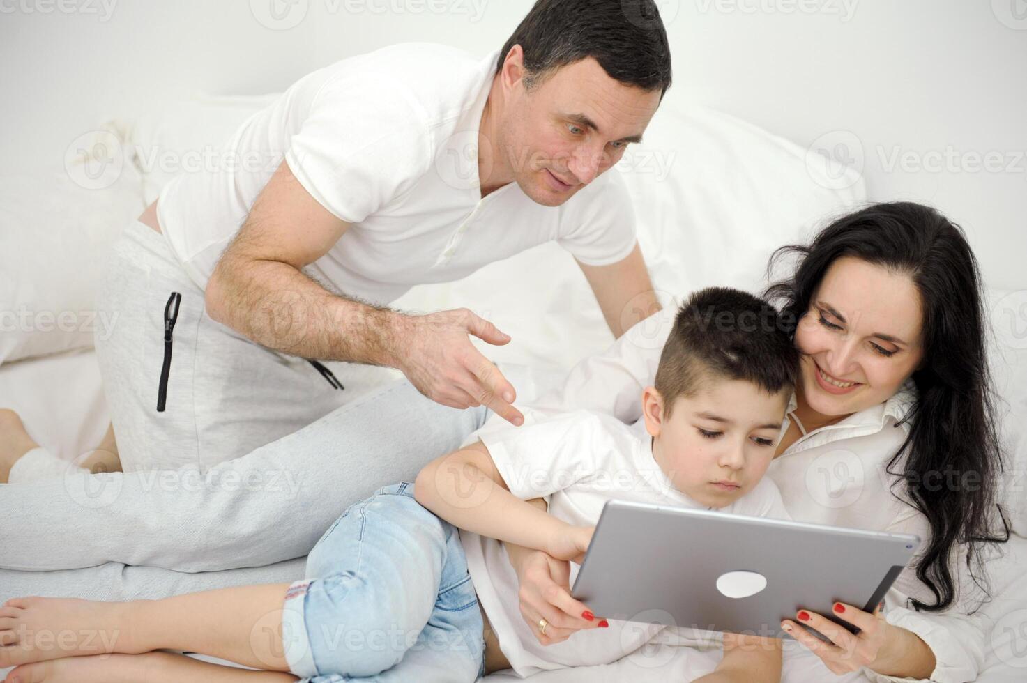 people, family and technology concept mother, father and little boy with tablet pc computer in bed at home or hotel room. Happy family spend time together lying under white blanket on bed at bedroom photo