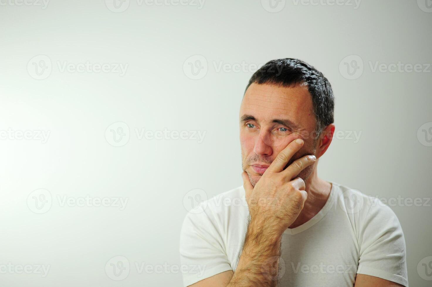 Doubt adult dark-haired middle-aged man is sad he put hand to chin put head on eyes when closed he thinks dreams reflects plans he has white t-shirt there is a place for text advertising blue eyes photo